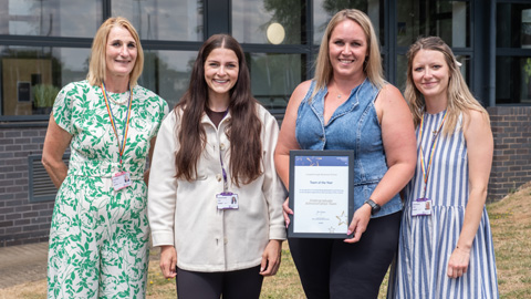 Three people standing in front of a building with large windows; one holding a framed certificate